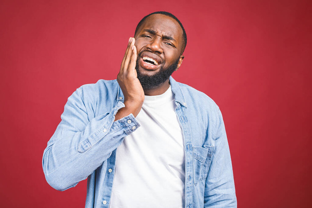 Young black man isolated on red background with mouth open, touching his face with expression of horrible suffer from health problem and aching tooth. - Photo, Image