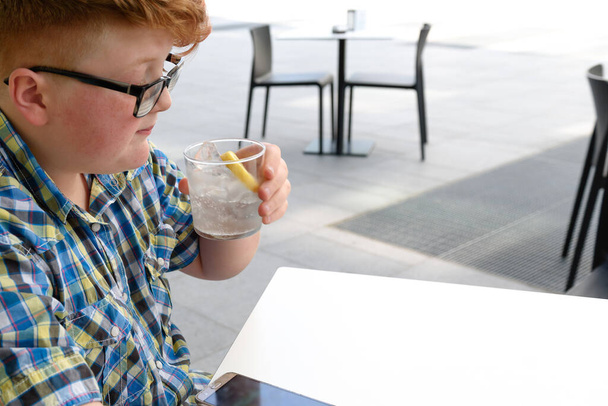 Rapaz ruivo com telemóvel a beber de um copo de água gelada com limão. Criança com óculos vestidos com uma camisa xadrez olha para o smartphone e bebe água
. - Foto, Imagem