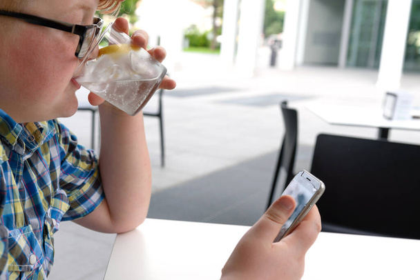Cute red-haired boy with cellphone and eyeglasses in an urban context drinks iced water with lemon. The boy with glasses looks at the screen of a smartphone and drinks water. - Photo, Image