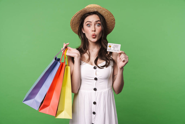 Image of amusing pretty woman in straw hat holding credit card and shopping bags isolated over green background - Фото, изображение