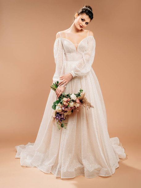 Wedding inspiration. Beautiful caucasian model in white wedding dress with decolletage and long sleeves holding trendy bouquet with fresh greenery.Full-length portrait of young smiling bride in studio - 写真・画像