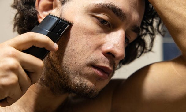 man shaving by trimmer morning action process view of adult person close face portrait in bath room - Photo, Image