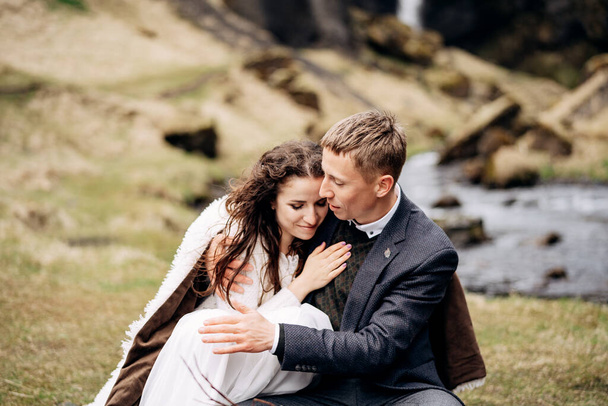 Mariage Destination Islande, près de la cascade de Kvernufoss. Un couple de mariés est assis sur les rives d'une rivière de montagne, à une table pour un dîner de mariage, faite d'une palette en bois peinte avec de la mousse et des chicots
 - Photo, image