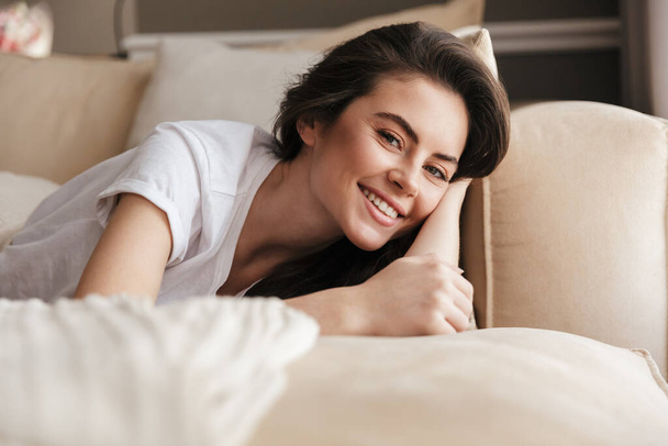 Beautiful smiling young brunette woman relaxing on a couch at home, looking at camera - Фото, изображение