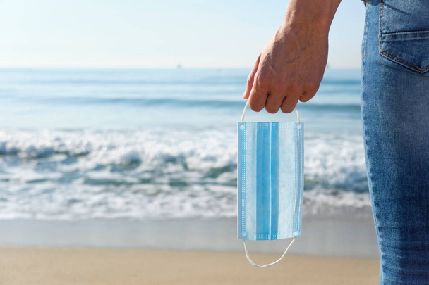 closeup of a young caucasian man in jeans, seen from behind, standing on the beach holding a blue surgical mask in his hand, as is taking a breather of wearing it, with the sea in the background - Photo, Image