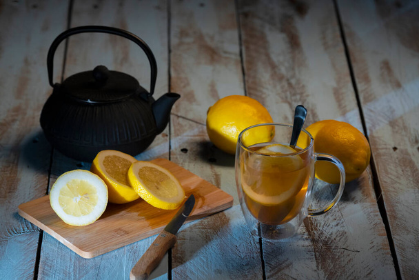 Planche à découper avec un citron tranché et un couteau entouré d'une tasse de thé, quelques citrons entiers et une théière noire sur une table en bois. Concept de détente et de style de vie
. - Photo, image
