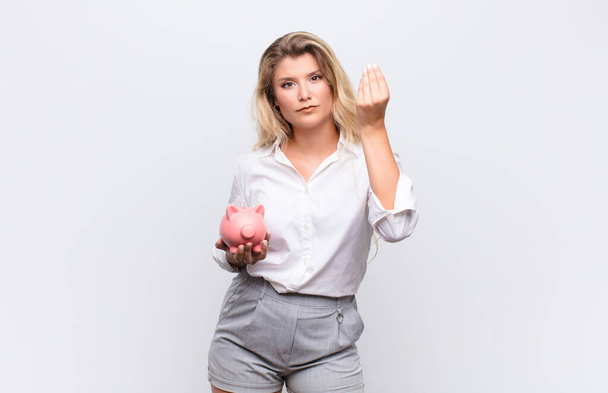 young pretty latin woman making capice or money gesture, telling you to pay your debts! with a piggybank against white wall - Fotoğraf, Görsel