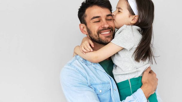 Indoor image of happy father smiling broadly hold embrace his cute daughter kissing on the cheek. Loving daddy and his little girl cuddling and enjoying time together. Childhood and fatherhood concept - 写真・画像