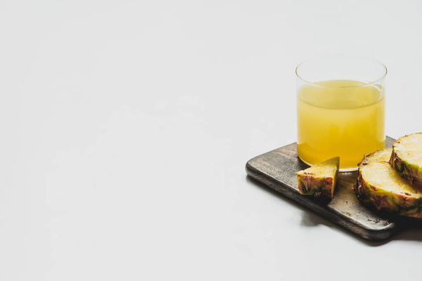 suco de abacaxi fresco em vidro perto de cortar frutas deliciosas em tábua de corte de madeira no fundo branco
 - Foto, Imagem