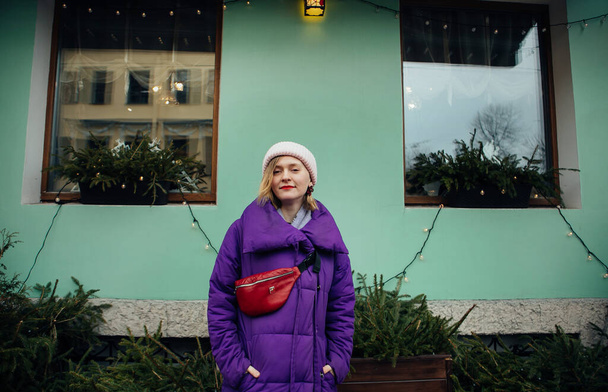 positive blonde in a purple down jacket and with a red belt bag walks around St. Petersburg - Fotografie, Obrázek