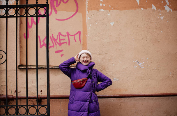 positive blonde in a purple down jacket and with a red belt bag walks around St. Petersburg - Foto, Imagen