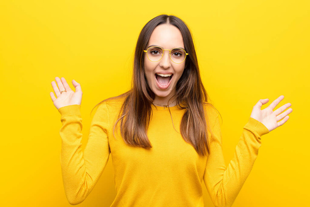 young pretty woman looking happy and excited, shocked with an unexpected surprise with both hands open next to face against orange wall - Fotoğraf, Görsel