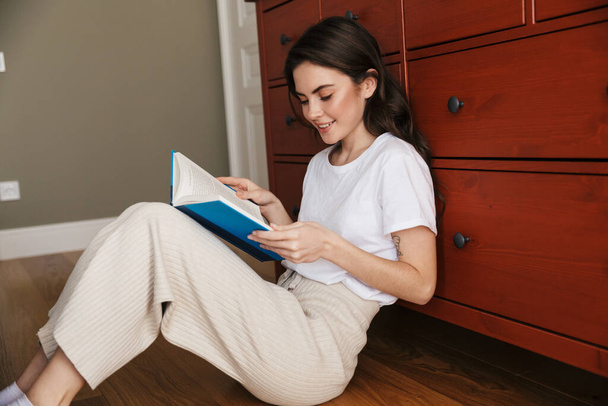 Attraente giovane donna bruna sorridente che legge un libro mentre si siede su un pavimento e si appoggia su un armadio
 - Foto, immagini