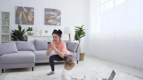Joyful pretty sporty young woman in sportswear using mobile during her fitness squating exercises while her small nice daughter playing on the carpet - Кадры, видео