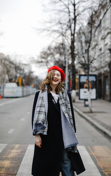 colorful blonde girl in a red hat, coat and scarf on a walk in the city smiling - Zdjęcie, obraz