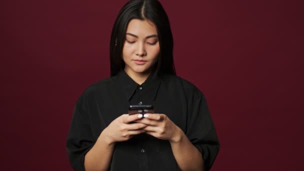 A pretty young asian woman wearing casual clothes focused on her phone then waving her hand making hello gesture isolated over red background - Filmagem, Vídeo