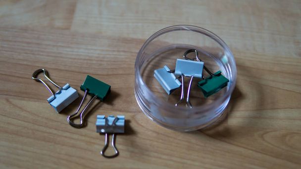 Top view of full binder clips in a transparent box and scattered on wooden background. It is a simple device for binding sheets of paper together. Selective focus on foreground. - Photo, Image