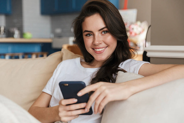 Beautiful smiling young brunette woman relaxing on a couch at home, using mobile phone - Foto, imagen