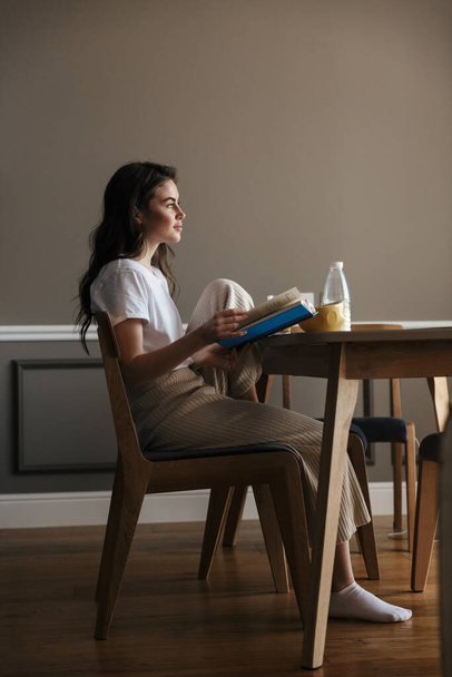 Attractive young brunette woman having a healthy breakfast reading a book while sitting at the kitchen table - Φωτογραφία, εικόνα