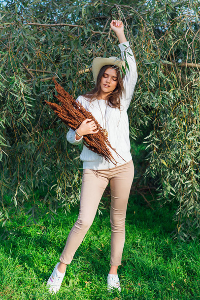 Happy beautiful brunette teenage girl dressed in a white sweater, jeans and cowboy straw hat standing behind tree brunches with green leaves holding dry bouquet - Foto, Imagen