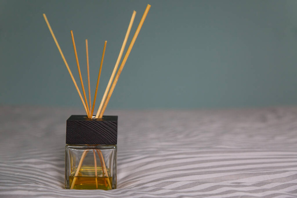a glass diffuser with a black wooden cover, wooden sticks and yellow aromatic oil stand on a gray bedspread. gray-blue wall in the background. selective focus - Photo, Image