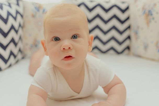 blue-eyed baby lies on his stomach in bed and looks surprised - Foto, Imagen