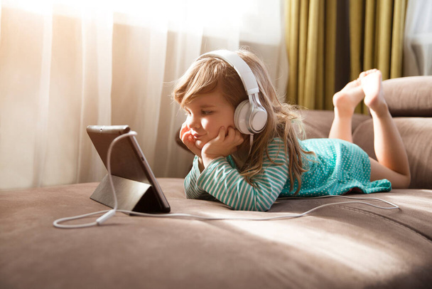 Child girl in headphones is using a digital tablet while lying on the sofa at home. Training online. quarantine. Corona Virus.  - Photo, Image
