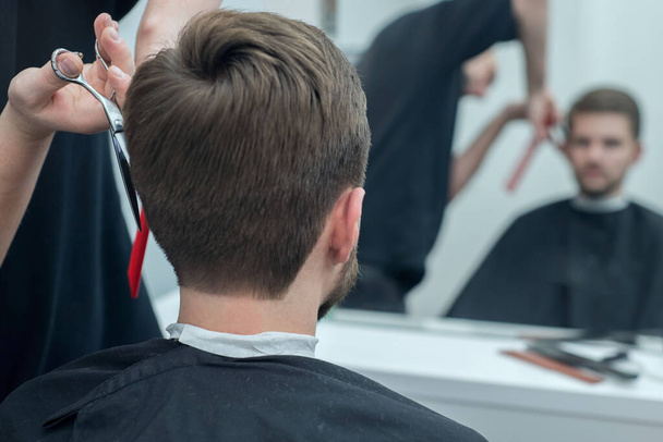Friseurmeister schneidet einem jungen bärtigen Mann die Haare auf den Kopf. Nahaufnahme der Hand eines professionellen Friseurs mit der Schere in einem Friseursalon. Männerfrisur. Frisur, Porträt eines bärtigen Typen beim Friseur. professionelle Haarpflege für Männer - Foto, Bild