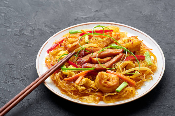 Singapore Mei Fun σε λευκό πιάτο σε σκούρο φόντο σχιστόλιθου. Singapore Noodles είναι κινέζικο πιάτο κουζίνας με χυλοπίτες ρυζιού, γαρίδες, char siu χοιρινό, καρότο, κόκκινο κρεμμύδι, λάχανο νάπα. Κινέζικο φαγητό. - Φωτογραφία, εικόνα