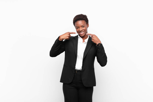 young pretty black womansmiling confidently pointing to own broad smile, positive, relaxed, satisfied attitude against white wall - Foto, immagini
