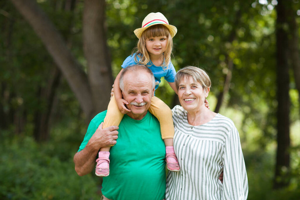 Grandparents giving  granddaughter ride on shoulders in summer outdoors. Concept of friendly family. - Photo, Image