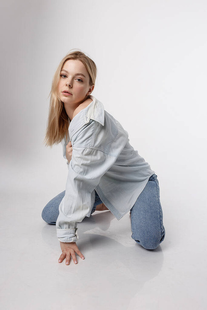 portrait of sexy caucasian woman with long hair posing in blue shirt and jeans on white studio background. model tests of pretty girl in basic clothes. attractive female sitting on floor on her knees - 写真・画像