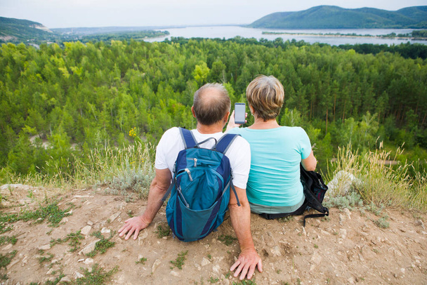elderly couple with backpacks sits on the mountain. Senior couple walking in nature. travel tourism concept. Social Distancing. end of a quarantine. Staycations, hyper-local travel,  family outing, getaway, natural environ - Foto, immagini