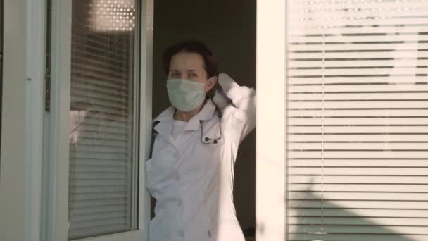 Zmęczona lekarka w kombinezonie ochronnym patrzy przez okno, zdejmuje maskę medyczną, zamyka oczy przed zmęczeniem, oddycha świeżym powietrzem. Ciężki dzień pracy lekarza w szpitalu. Pandemiczny koronawirus - Materiał filmowy, wideo