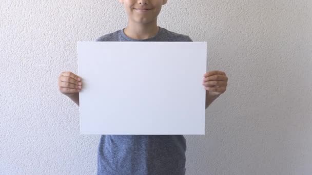 Chlapec drží v rukou a ukazuje prázdný bílý plakát. Šedá stěna na pozadí. Zavřít - Záběry, video