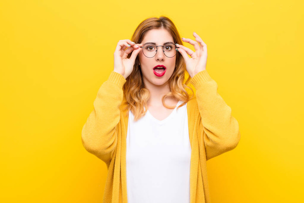 young pretty blonde woman feeling shocked, amazed and surprised, holding glasses with astonished, disbelieving look against yellow wall - Foto, Bild