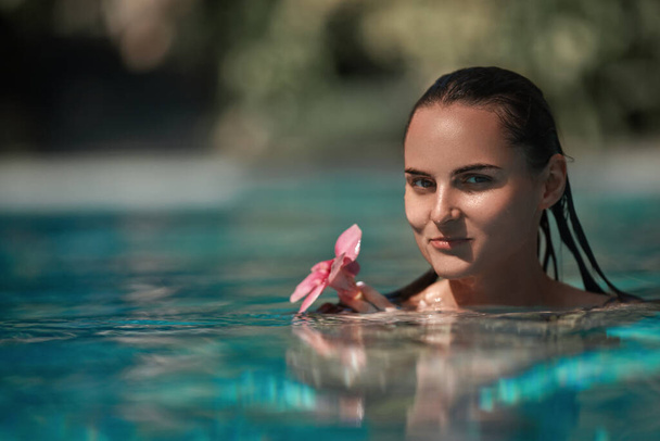 Portret pięknej brunetki z mokrymi włosami pływającej w basenie i trzymającej egzotyczny purpurowy kwiat w rękach. Pojęcie podróży, relaksu i przyjemności - Zdjęcie, obraz
