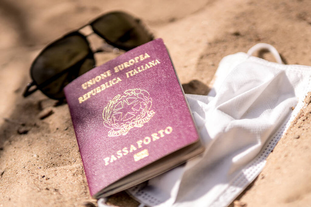 Podróżował podczas epidemii koronawirusa. Okulary przeciwsłoneczne, maska ochronna i włoski paszport na piaszczystej plaży - Zdjęcie, obraz