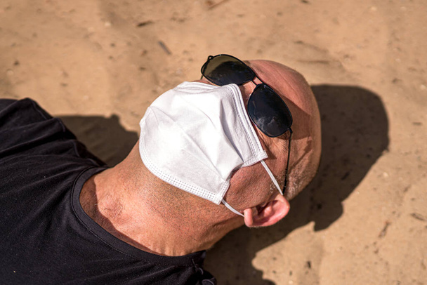 コロナウイルス感染症の発生中に日光浴.ロックダウンやコロナウイルス病の発生中の夏休み。砂浜でサングラスと医療用保護マスク日光浴を身に着けている男 - 写真・画像