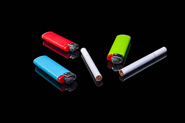 witte filtersigaretten en plastic gasaanstekers van verschillende kleuren op een geïsoleerde zwarte achtergrond met reflectie - Foto, afbeelding