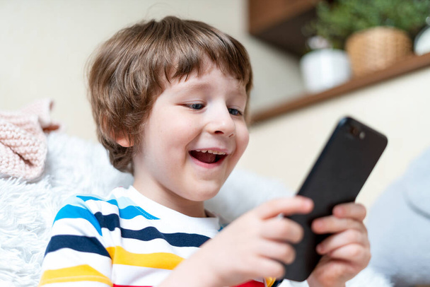 Счастливый маленький мальчик играет в онлайн-игру, смотрит видео по мобильному телефону, сидит на диване и развлекается в гостиной. Улыбает малыша с помощью смешных мобильных приложений, наслаждаясь свободным временем дома. - Фото, изображение