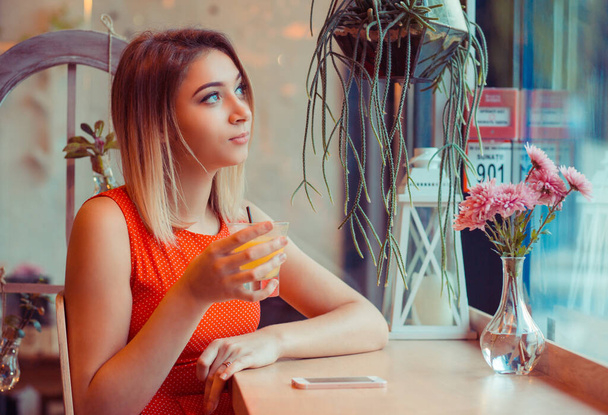 Cafe city lifestyle thoughtful woman drinking orange juice, looking outside on street through glass window, sitting indoor juice bar, in home kitchen or in coffee shop, trendy cafe wearing red dress - 写真・画像