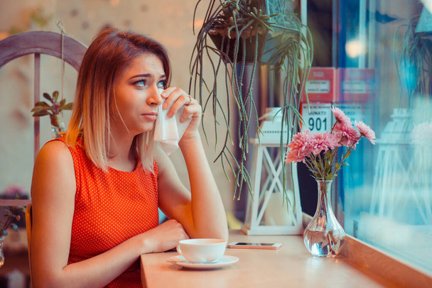 Niedola, infekcja. Brunetka dziewczyna płacze wycierając łzy kichając w chusteczce pijąc herbatę w swojej kuchni, w pobliżu okna lub kawiarni, modna kawiarnia w czerwonej sukience. Negatywne emocje twarzy - Zdjęcie, obraz