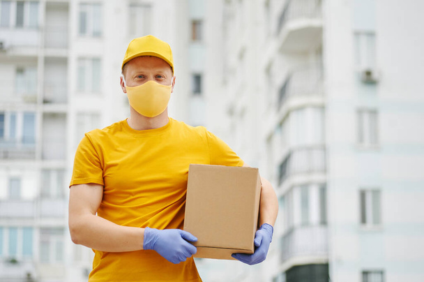 Teslimatçı eleman sarı üniforma şapkası, tişört, yüz maskesi ve eldiven içinde bina zemininde karton bir paket tutuyor. Covid-19 virüs salgınında güvenlik karantina hizmeti. - Fotoğraf, Görsel