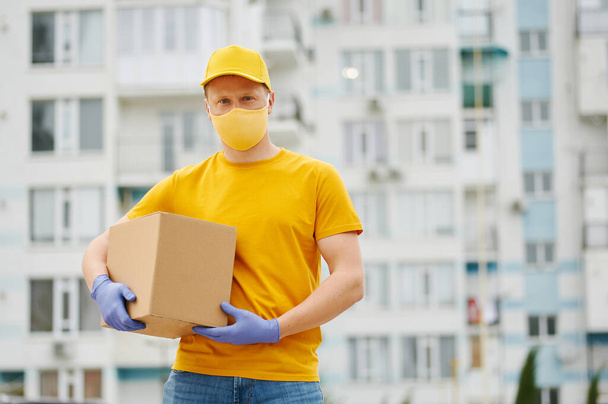Delivery Man υπάλληλος σε κίτρινο ομοιόμορφο καπέλο, t-shirt, μάσκα προσώπου και γάντια κατέχει ένα πακέτο κουτί από χαρτόνι για την οικοδόμηση φόντο. Υπηρεσία καραντίνας κατά την περίοδο πανδημίας του ιού covid-19. - Φωτογραφία, εικόνα