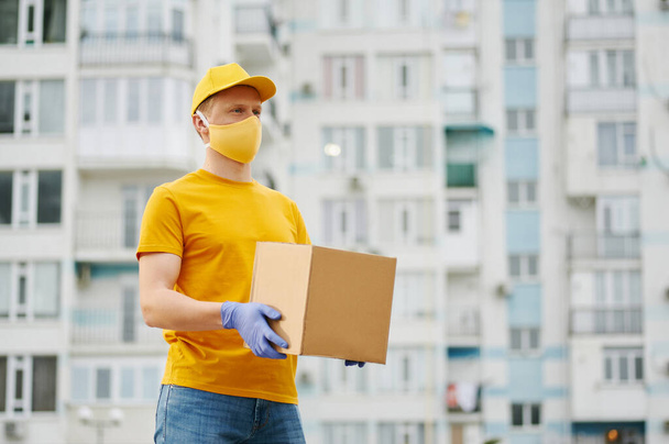Delivery Man worker in yellow uniform cap, t-shirt, face mask and gloves holds a cardboard box package on building backdrop. Safety delivery quarantine service in covid-19 virus pandemic period. - Photo, Image