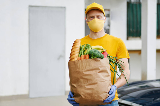 Grocery Delivery Man in yellow uniform cap, t-shirt, face mask, gloves holds a paper bag with food, fruit, vegetables on building backdrop. Quarantine delivery service in covid-19 virus pandemic. - 写真・画像