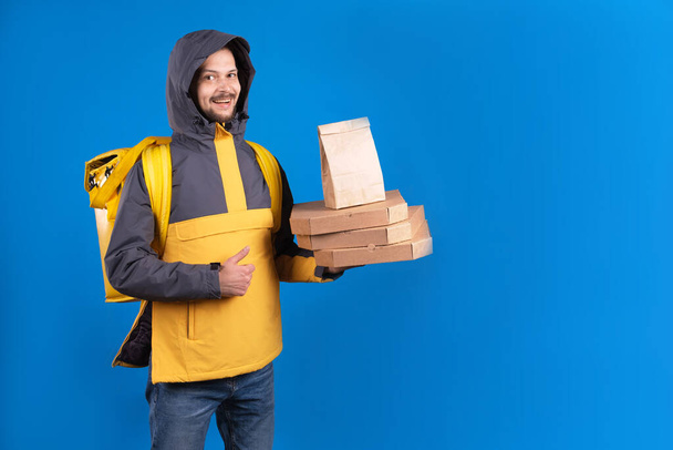 Unshaven dark-haired caucasian courier in yellow windbreaker holds an order of pizza and coffee and refrigerator bag for food, delivery man shows an approving hand gesture symbolizing good delivery - Valokuva, kuva