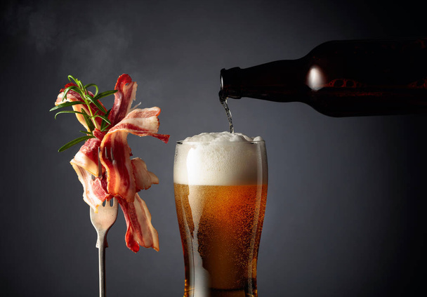 Beer and slices of fried bacon garnished with rosemary. Beer is poured from a bottle into a glass.  - Foto, afbeelding