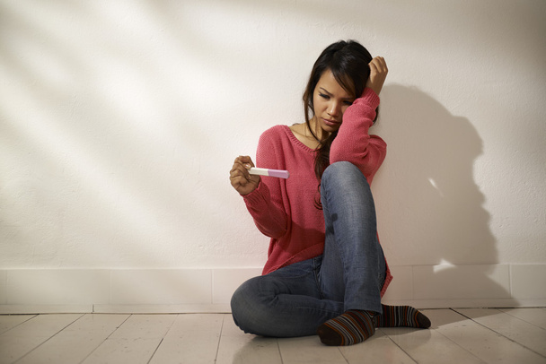 Грустная азиатка смотрит на тест на беременность сидя на полу
 - Фото, изображение